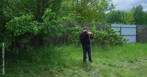 A child preserving the overgrown lawn in the backyard, using a gasoline trimmer, near a wooden fence and trees, performing seasonal work in the yard during a sunny summer day.