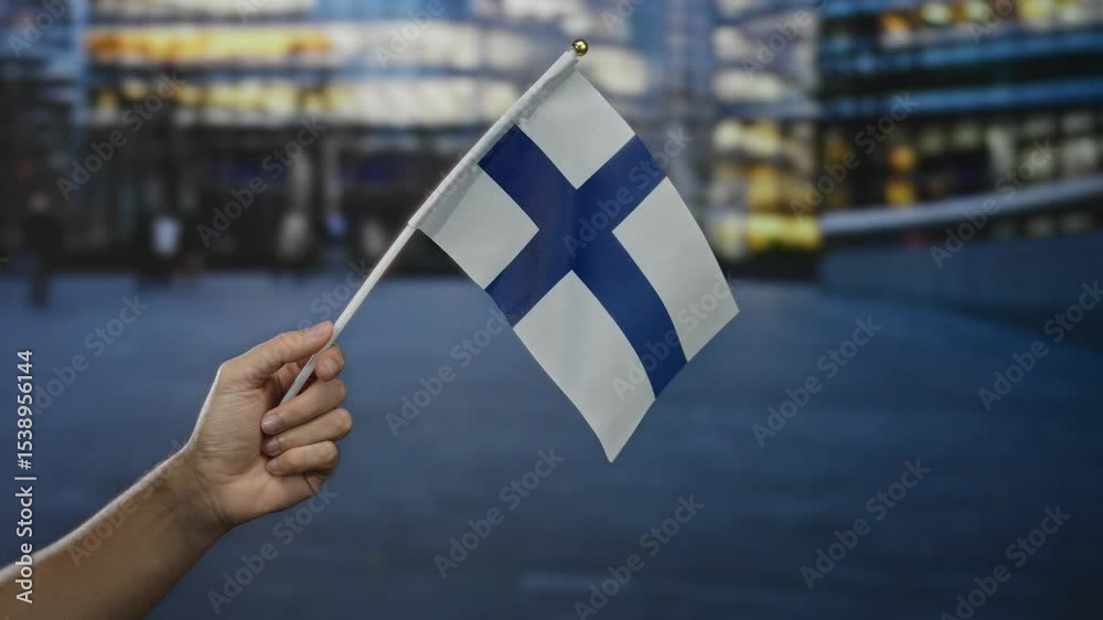 Hand holding finland flag in city street scene with blurred urban background during dusk representing national pride and culture.