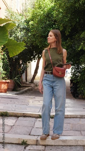Young woman walks down stone steps in the old town of Athens, Greece. She wears a green top and jeans, holding a phone. Sunny day, lush plants, casual moment. Vertical video