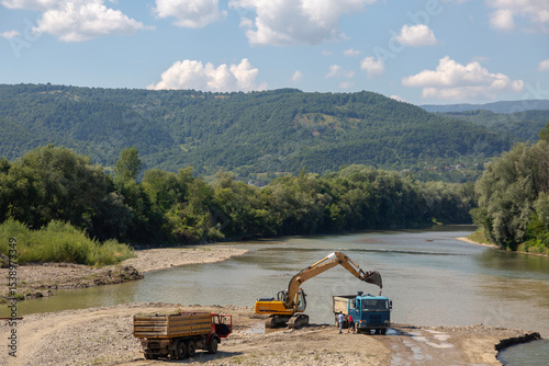 Industrial activity along a river bank: an excavator loads material into trucks, set against a backdrop of rolling forested hills
