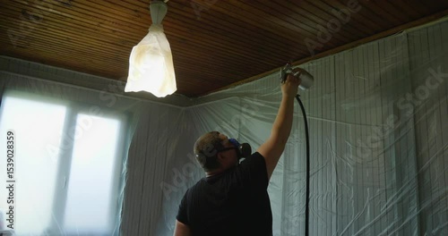 A man in a protective mask uses an aerosol gun, paints a wooden ceiling in white, with plastic sheets covering the walls to protect them from spray of paint.