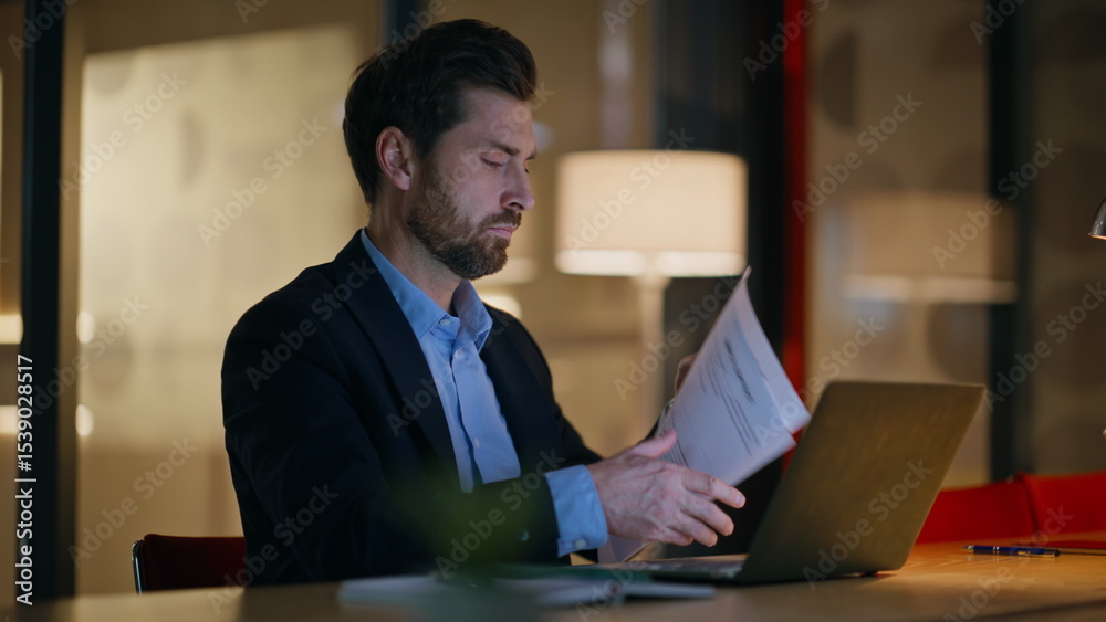 Obraz premium Late worker reading papers working at laptop in dark office closeup. Serious man