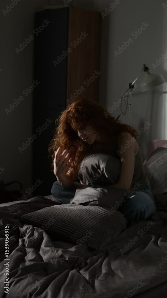 Vertical shot of red-haired woman curled on bed feeling distressed and angry in dimly lit room