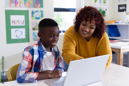 Female teacher helping african american boy with laptop in classroom, both smiling, at school photo