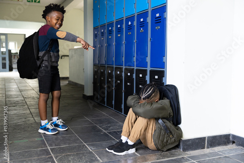 In school hallway, boy teasing another sitting by lockers, showing bullying