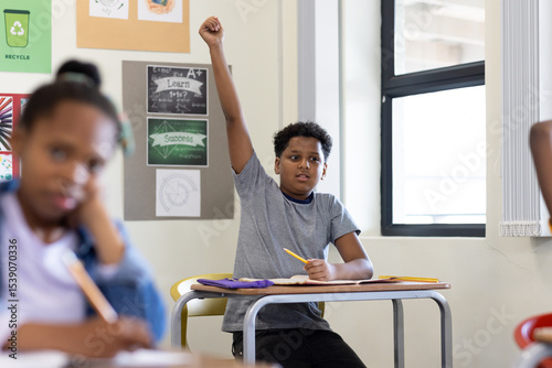 In school, boy raising hand eagerly during classroom lesson, holding pencil