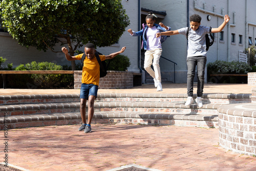 Joyful diverse students with backpacks jumping off steps outside school building