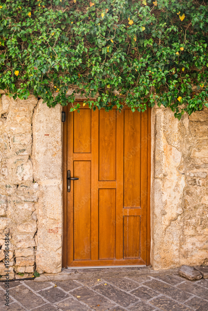 Fototapeta premium Wooden door framed by ivy and ancient stone wall. Summer travel, rustic charm, and the warmth of Mediterranean architecture in small historic towns.