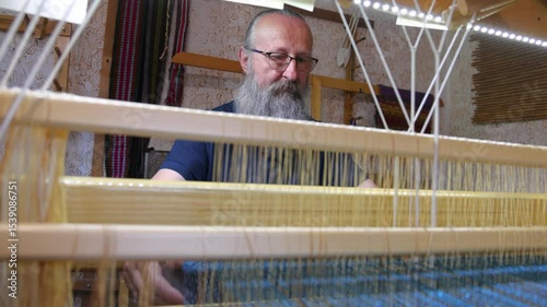 Weaver at the traditional loom. Mature man with a graying beard and glasses is weaving cloth and tapestry on a loom, using traditional techniques handicraft in the production of textile garments