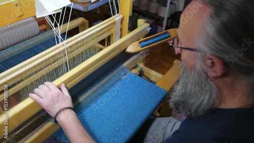 Traditional weaver at the loom. Mature man with a graying beard and glasses using a weaving shuttle works at the loom, weaving cloth and tapestry by hand in textile garment production