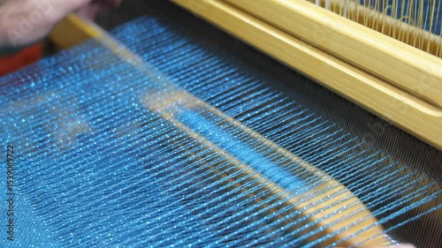 Weaver at the traditional loom. Mature man using a weaving shuttle works at the loom, weaving cloth and tapestry by hand as part of traditional techniques in textile garment production