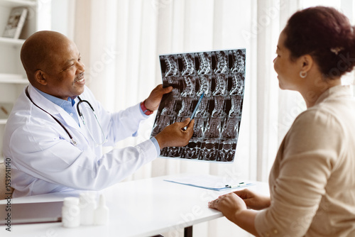 Smiling African American male doctor in medical coat explaining spine x-ray results to attentive black female millennial patient during appointment in modern clinic, closeup