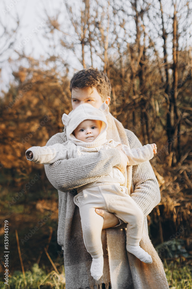 Fototapeta premium Happy father hugging cute baby son in autumn field in soft evening light. Parenthood. Portrait of dad and little child in cozy stylish clothes embracing in fall park