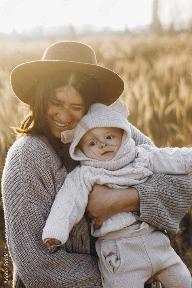 Obraz premium Happy mother hugging cute baby son in autumn field in soft evening light. Parenthood. Mom and little child in cozy stylish clothes embracing in fall park, love tranquil moment