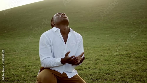 African-American man kneeling in prayer on a green field, wearing a white shirt.  
