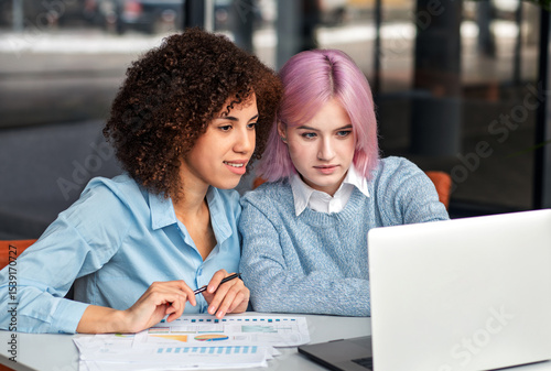 Young diverse women working together at laptop and analyzing business documents. Concept of teamwork, digital collaboration, mentorship and modern workplace