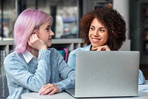 Smiling diverse young women collaborating on laptop in modern coworking space. Concept of teamwork, digital nomads, freelance, generation Z and creative business