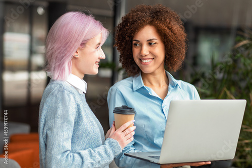 Two smiling young women having friendly conversation while holding laptop and coffee in modern workspace. Concept of networking, teamwork and female empowerment