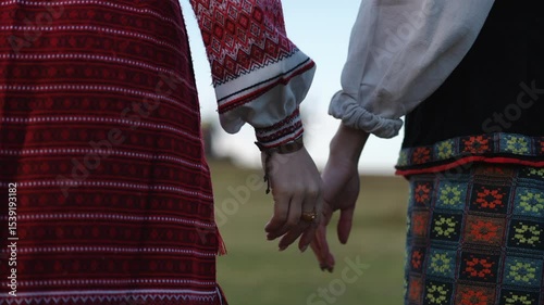 Close-up of two women wearing traditional embroidered garments, standing side by side and holding hands in a field. Their gesture expresses tenderness and connection, with focus on the details of the