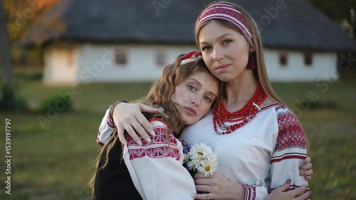 Two girls in traditional embroidered clothing share a gentle embrace outdoors. One leans her head on the other's shoulder, holding a bunch of field flowers.