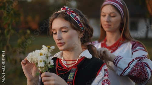 One girl carefully braids her friend's hair as the other gently holds a bouquet of daisies, both wearing traditional folk outfits in a calm rural evening.