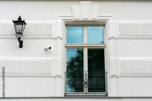 Modern white building facade with classic decorative elements, vintage street lamp, security CCTV camera and window with reflection and metal balcony railing on a sunny day. Urban architecture detail.