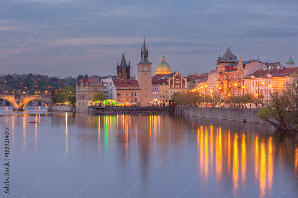Naklejka premium Colorful buildings and towers along the Vltava River in Prague Czech Republic at sunset with calm water and vibrant sky