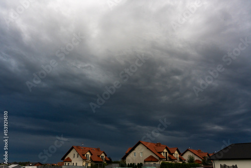 Dramatic storm clouds gathering over suburban houses with red roofs in residential area on dark evening. Concept of extreme weather, thunderstorm approaching and atmospheric tension.