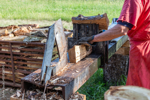 Lumberjack wearing gloves is splitting firewood logs with a hydraulic machine in a rural countryside setting, surrounded by a pile of already split logs in the background