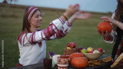 A cheerful woman in traditional Eastern European clothing hands a pumpkin to another person at a rustic outdoor table decorated with autumn fruit and pottery.