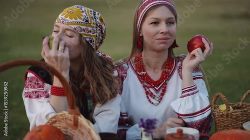 One woman in embroidered clothing holds a red apple near her lips while the other looks into a harvest basket filled with fruit and pumpkins on a rustic outdoor table.