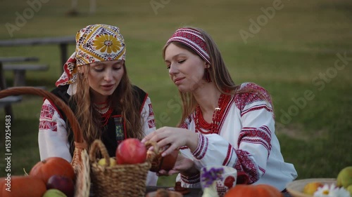 Two women in traditional Ukrainian clothing sit at a wooden table in the open air, holding warm mugs, surrounded by pumpkins, apples, and flowers on a calm autumn evening.