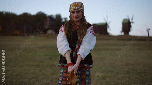 Girl in traditional Ukrainian outfit gently sways her shoulders while holding autumn leaves, windmills visible in the background under an evening sky.