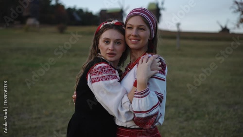 Two young women dressed in traditional embroidered outfits are dancing hand in hand in a wide open field. The moment captures lighthearted motion, harmony, and connection at sunset.