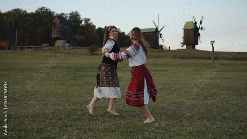 Joyful scene of two women spinning barefoot in folk costumes on a rural field. Their embroidered outfits flow naturally as they dance near wooden windmills in warm evening light.