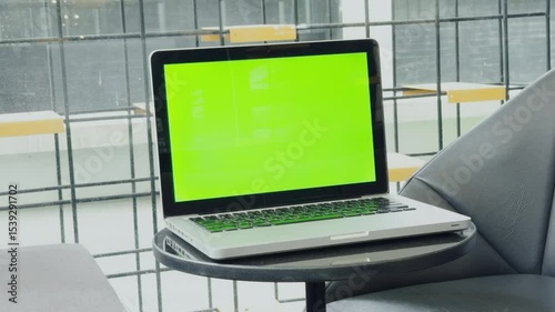 Laptop with green screen on a table in a bright office setting, featuring a scenic view through large windows.