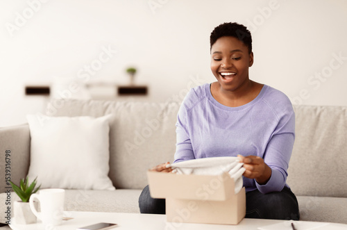 Delivery Concept. Smiling chubby black woman unboxing cardboard package, sitting on the couch in living room at home, free space. Excited lady happy with purchase and clothes shipment photo