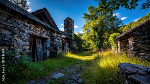 Overgrown stone chapel and outbuildings with bell tower in rustic, weathered condition under blue sky