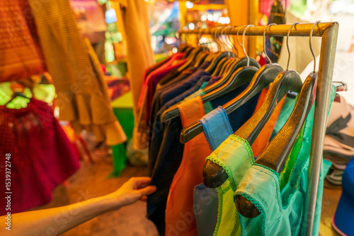 Fototapeta Naklejka Na Ścianę i Meble -  Woman shopping for colorful traditional clothes at rural Thai street market where local vendors sell handmade garments to tourists during cultural travel experience in countryside Thailand