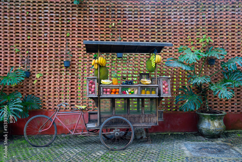 Vintage fruit cart with bananas, jackfruit, and other fruits stands against brick wall, surrounded by lush plants
