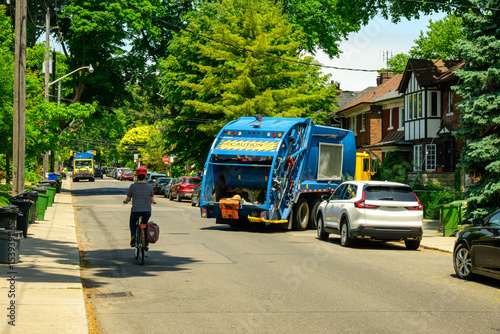 garbage trucks on the street in an older treelined residential neighbourhood with bins out for pick up with a cyclist and parked cars shot in summer in Toronto	