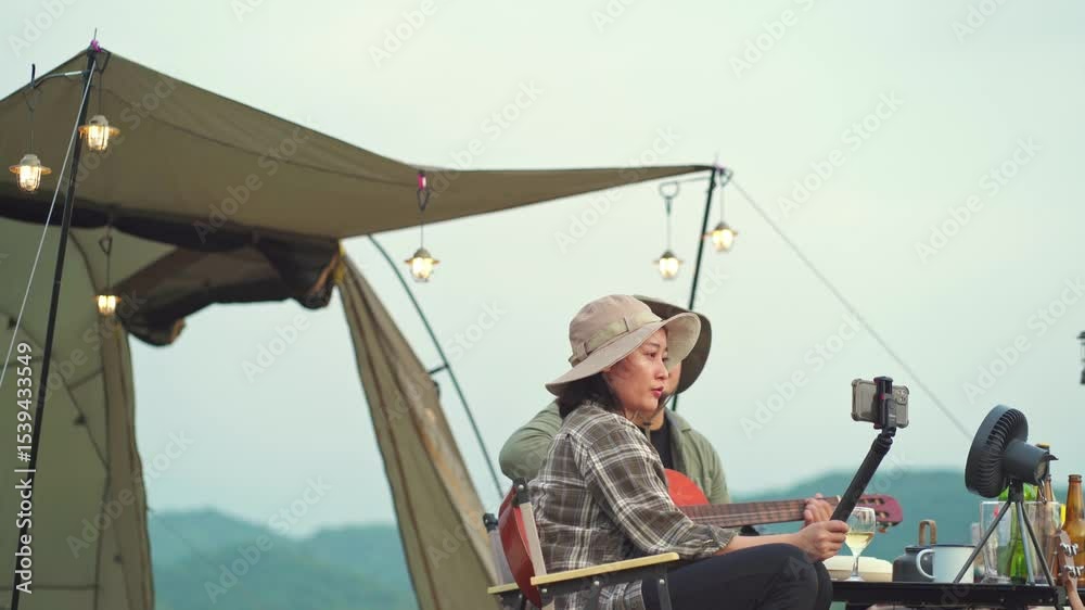 Asian couple camping on a grassland, woman smiling while taking a selfie with hands and man happily playing guitar, surrounded by nature, tranquility and outdoor happiness.