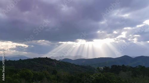 Sun rays breaking through cloudy sky over rolling hills of distant mountains covered in lush green forests and trees landscape