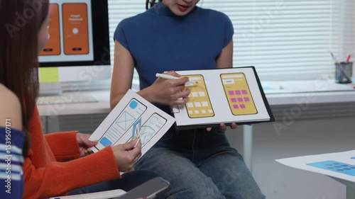 Web designers collaborating on mobile app layouts, comparing paper drafts with digital mockups on tablet during creative project planning meeting