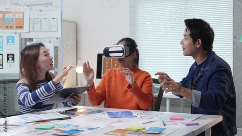 UI and UX designers testing virtual reality app in the office, woman wearing VR headset interacting with her colleague while sitting at table with tablet and app design sketches