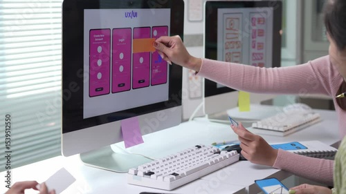 Two Asian women, probably user experience and user interface designers, are discussing and testing a new mobile app interface using sticky notes on a computer screen in an office