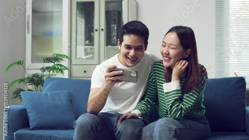 Young asian couple relaxing together on living room sofa, sharing smartphone content and enjoying hilarious moments while laughing with genuine connection