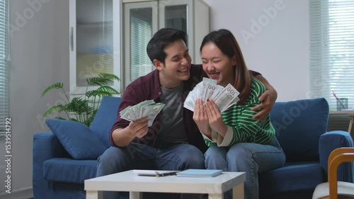 Happy asian couple sitting on home sofa, counting personal earnings and celebrating financial success with wide smiles and shared excitement about their economic achievements