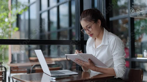 Professional Asian businesswoman concentrating on work, writing notes while using laptop at cafe table during daytime, capturing focused career moment with modern urban backdrop