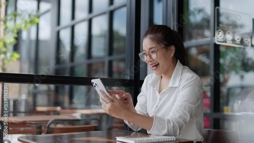 Joyful young professional sitting at table in cafe, receiving positive news on smartphone and celebrating career success with genuine excitement